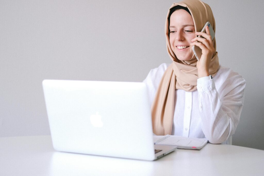 Smiling woman in a hijab uses laptop and phone at a home office.