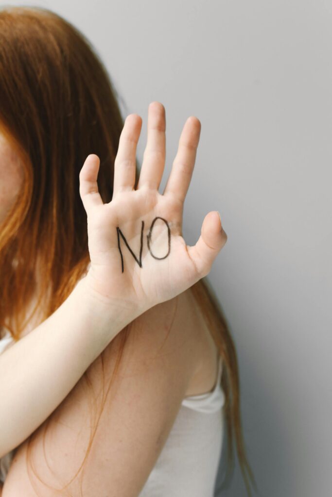 A woman with red hair raises her hand with 'No' written on her palm against a neutral background.