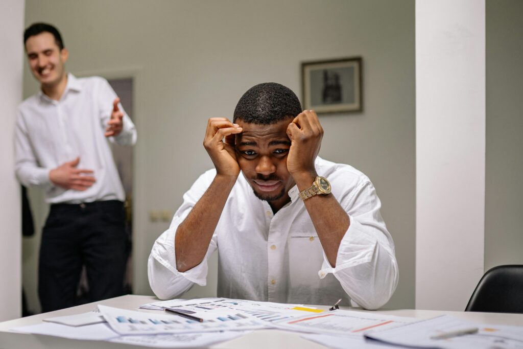 Tensed businessman at desk while coworker is laughing. Office stress concept.